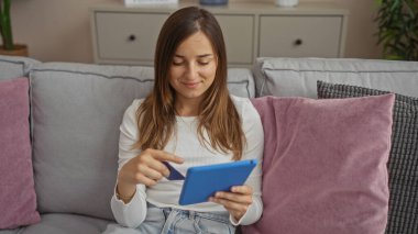Genç bir kadın oturma odasında elinde bir kredi kartıyla oturuyor ve rahat yastıklarla çevrili bir tablet kullanıyor..