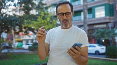 Şehir parkında cep telefonuyla meşgul, şehir manzarası ve arka planda ağaçlarla çevrili, şaşırdığını ifade eden olgun İspanyol bir adam.