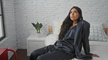 Genç, çekici bir kadın şık, modern bir yatak odasında beyaz tuğlalı duvarları ve iç dekorasyonu olan bir yatakta dinleniyor..