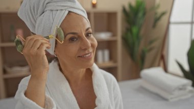 Orta yaşlı İspanyol bir kadın spa odasında yüzüne yeşim bir silindir uyguluyor, sakin bir ifadeyle beyaz bir bornoz giyiyor, sakin bir iç güzellikle çevrili..