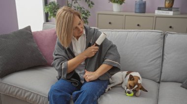 Orta yaşlı beyaz bir kadın, oturma odasında gri bir kanepede otururken ve yanında küçük bir köpek oynuyorken elbisesinde pamuk sarma kullanıyor..