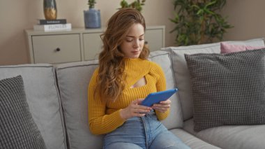 Sarışın, güzel bir kadın, rahat bir oturma odasında rahat bir koltukta oturuyor mavi bir tabletle nişanlanmış, şık sarı bir kazak ve kot pantolon giyiyor..