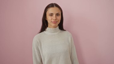 Genç, çekici, İspanyol bir kadın, izole edilmiş pembe bir arka plana güvenle gülümsüyor, zarif ve zarif bir ifade sergiliyor, açık renk bir süveter giyiyor.