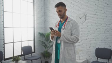 Sakallı İspanyol erkek doktor klinik bekleme odasında modern dekorlu akıllı telefon kullanıyor..