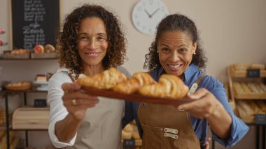 Women bakers smiling in bakery holding tray of freshly baked croissants in cheerful, inviting shop interior featuring bread shelves and wall clock in background clipart