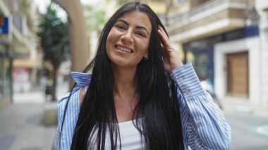 Şehir caddesinde gülümseyen esmer, genç ve çekici bir kadın uzun saçlarını açık havada günlük kıyafetleriyle sergiliyor..