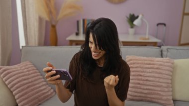 Kapalı alanda kutlama yapan genç bir kadın modern dekorlu ve pastel renkli rahat bir oturma odasında telefonuna bakıyor.