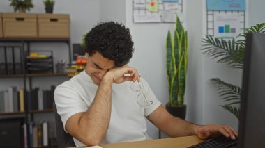 Kıvırcık saçlı genç, İspanyol bir adam ofis masasında oturuyor, elinde gözlüklerini tutuyor ve bitkin görünüyor..