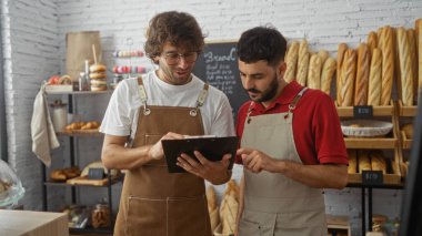 İki adam, biri kırmızı tişörtlü, biri beyaz gömlekli, önlüklü ve pastanede panoya bakan, arkasında ekmek ve hamur işi rafları olan iki adam..