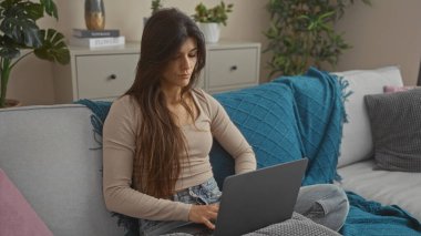 Genç bir İspanyol esmer kadın kanepede oturuyor. Rahat bir oturma odasında, üzerinde bitkiler ve mavi bir battaniye olan bir dizüstü bilgisayar kullanıyor..