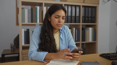 Ofis ortamında okurken, arka planda kitap rafları ve dosyalarla akıllı telefonda yazan genç bir kadın..