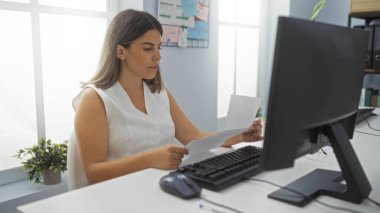 Ofis masasında bilgisayarlı ve bitkili evrakları inceleyen bir kadın, iş sırasında kapalı bir yerde, parlak bir iş yerinde odaklanmış görünüyor..