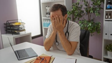 Stresli İspanyol erkek doktor hastane ofisinde masasında oturuyor. Yüzü elleri tıbbi malzemelerle çevrili..