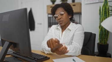 Ofisteki bir bilgisayarda çalışan bir kadın, arka planda iç mekan bitkileri ve ofis dekorlarıyla çevrili kıvırcık saçları ve gözlükleriyle rahatsız olduğunu belirterek bileğini tutuyor.