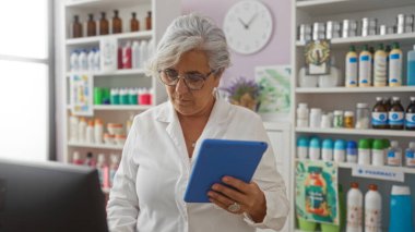 Yaşlı kadın, gri saçlı, eczanede duruyor, mavi bir tabletten okumaya odaklanmış, ilaç ürünleri ve çeşitli sağlık ürünleriyle dolu raflarla çevrili..