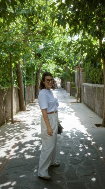 Kapri, İtalya 'da güneşli bir günde, güzel bir Avrupa caddesi boyunca yemyeşil bir çevre ile çevrili, neşeli bir cazibe ve gençlik dolu bir kadın..