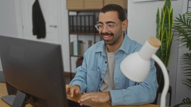 Ofiste çalışan İspanyol bir adam, bilgisayarda yazı yazıyor ve elinde lambayla bir masada otururken gülümsüyor. Etrafı kitaplık ve bitkilerle çevrili..