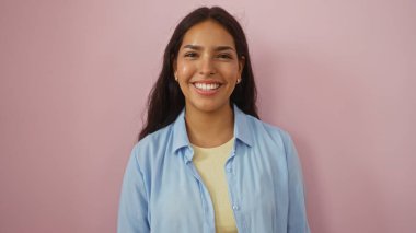 İzole edilmiş pembe arka planda duran gülümseyen genç bir kadın. Günlük açık mavi gömlek ve beyaz atlet giyiyor.