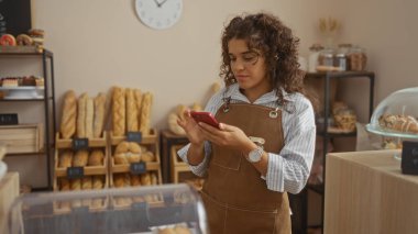 Etrafı ekmek ve hamur işleriyle çevriliyken fırında akıllı telefon kullanan genç bir kadın..