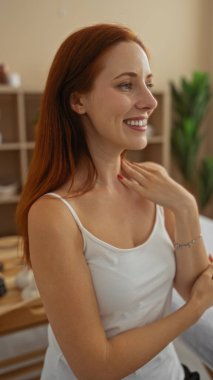 Young woman with red hair smiling in an indoor wellness spa setting while wearing a white tank top clipart