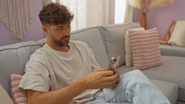 Kısa kıvırcık saçlı genç adam gri bir kanepede oturmuş pembe yastıklı ve dekoratif unsurları olan rahat bir oturma odasında akıllı telefon kullanıyor.