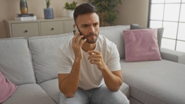 Oturma odasında oturan ve telefonda konuşan sakallı İspanyol genç adam endişeli görünüyor..