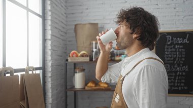 Fırında içki içen genç bir adam, önlük giymiş, etrafı ekmek ve hamur işleriyle çevrili.