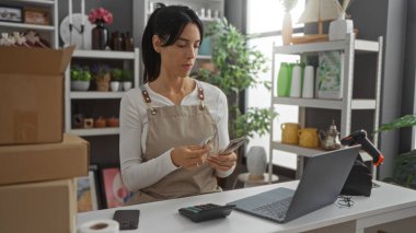 Ev dekorasyonu mağazasında para sayan bir kadın dizüstü bilgisayar kullanıyor. Etrafı bitkiler ve ev aksesuarlarıyla çevrili.