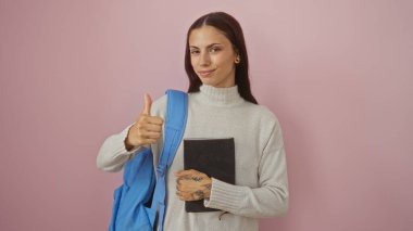 Mavi bir sırt çantası ve not defteri olan genç bir kadın pembe arka plan duvarına başparmak işareti yapıyor. Pozitif tavır ve kendine güven sergiliyor..