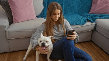 Beyaz köpeği yerde onun yanında otururken genç sarışın bir kadın oturma odasında telefonunu kullanıyor..