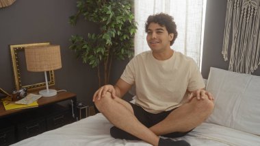 Latin kökenli genç bir adam yatak odasındaki rahat bir yatakta bağdaş kurarak oturur. Etrafı lamba, çerçeveli resimler ve bir bitkiyle çevrilidir..