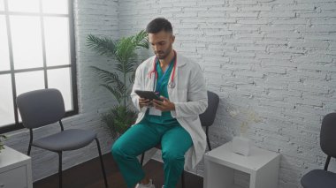 Sakallı genç İspanyol erkek doktor klinik bekleme odasında tablet kullanarak oturuyor..