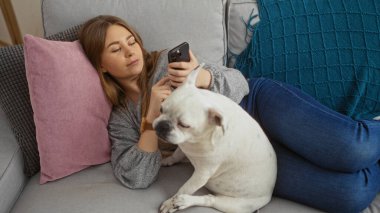 Genç sarışın bir kadın oturma odasındaki kanepede dinleniyor telefonu ve beyaz köpeğiyle..