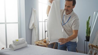 Klinikte bir adam havlularla masaj yatakları hazırlıyor, temiz ve sakinleştirici bir terapi ortamı öneriyor, deneyimli bir profesyonelin titiz ayarlarını vurguluyor.