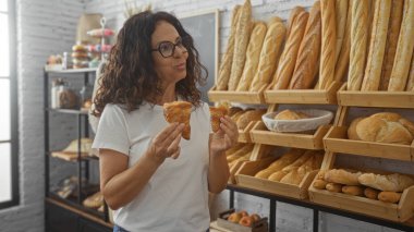 Taze ekmek ve hamur işleriyle çevrili bir pastanede kruvasan yiyen, gözlüklü ve beyaz gömlekli bir kadın, mutluluğu gösteriyor ve evin içinde anın tadını çıkarıyor..