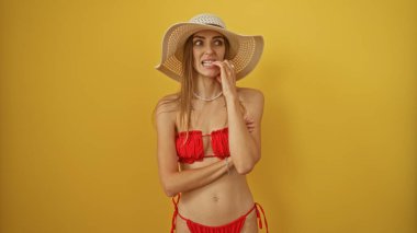 Kırmızı bikinili güzel sarışın kadın gergin bir şekilde sarı arka planda tırnaklarını yiyor.