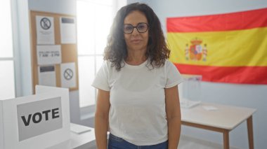 Bir kadın elinde İspanyol bayrağıyla kapalı bir odada duruyor. Arka planda pankartlar, pankartlar ve oy sandığıyla seçim temasını gösteriyor..