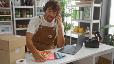 Genç adam, tasarım ürünleri ve dizüstü bilgisayarla çevrili bir şekilde telefonla konuşurken bir ev dekorasyonu dükkanında çalışıyor..