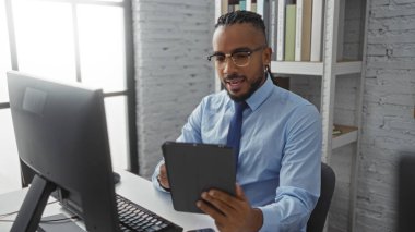 Örgülü genç adam modern ofis ortamında tablet kullanıyor, gözlük takıyor ve mavi gömlek giyiyor, çağdaş iç mekan ve teknoloji unsurları ile çevrili, odak ve bağlılık gösteriyor..