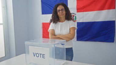 Seçim kolejindeki İspanyol kadın oy sandığı ve Dominik Cumhuriyeti bayrağı önünde kollarını kavuşturdu..