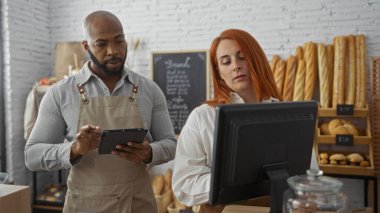 Erkek ve kadın fırının içinde birlikte çalışıyor, taze ekmek ve raflarla çevrili bir tablet ve bilgisayara odaklanıyorlar.