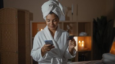 Bornoz ve havlu giymiş İspanyol bir kadın, arka planda mumlarla loş bir sağlık odasında kredi kartı tutarken cep telefonu kullanıyor..