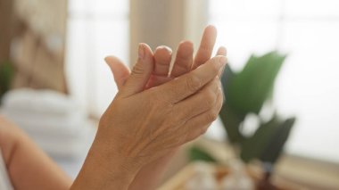 Yaşlı bir kadın, arka planda bulanıklık olan bir spa odasında ellerini uzatıyor. Bu da içeride huzurlu ve rahatlatıcı bir sağlık deneyimi olduğunu gösteriyor..