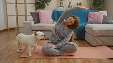 Sarışın genç bir kadın, rahat bir evde beyaz köpeğiyle yoga yapıyor. Pembe bir paspasın üzerinde, rahat bir dekor ve bitkilerle çevrili..