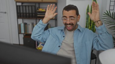 Genç İspanyol adam modern ofis ortamında arka planda kitaplıklarla başarıyı kutluyor.