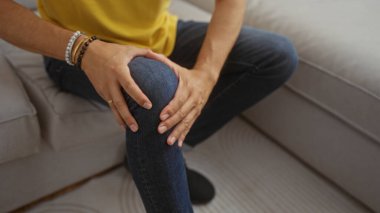 Sarı tişörtlü bir adam evde bir koltukta oturuyor dizini iki eliyle tutuyor, bileğinde bir bilezik var..