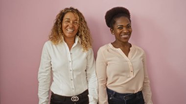 İzole edilmiş pembe bir arka planda duran kadınlar, dostluk ve çeşitlilik bağlarını sergiliyorlar, biri Afro-Amerikan, diğeri ise Latin Amerikalı, ikisi de gömlek ve kot pantolon içinde gülümsüyor..