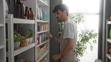 Sakallı yakışıklı İspanyol bir adam mağazadaki dekoratif ürünlerle dolu iç mekan bitkileri ve raflarla çevrili dekoratif eşyalara bakıyor.