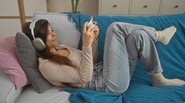 Esmer saçlı genç bir İspanyol kadın modern bir apartman dairesinde dinlenirken kulaklıkla müzik dinliyor. Kanepede uzanıyor ve telefonunu kullanıyor..