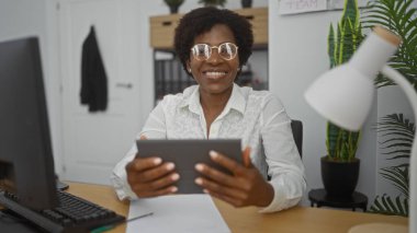 Kıvırcık saçlı, gülümseyen Afro-Amerikan kadın ofis ortamında çalışıyor, tablet kullanıyor, ofis malzemeleri ve kapalı bitkilerle çevrili..